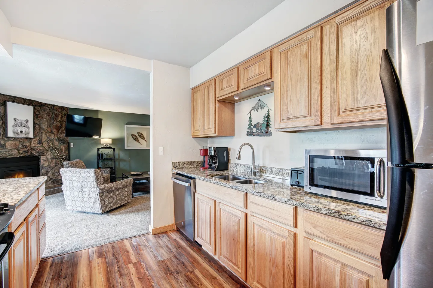 Kitchen with wood cabinets and granite-look countertops leading into a living room with a stone fireplace.