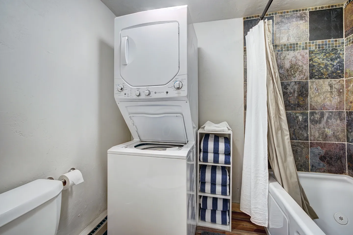 Bathroom with a stacked washer and dryer, toilet, towel shelving unit, and bathtub with tiled wall surround.