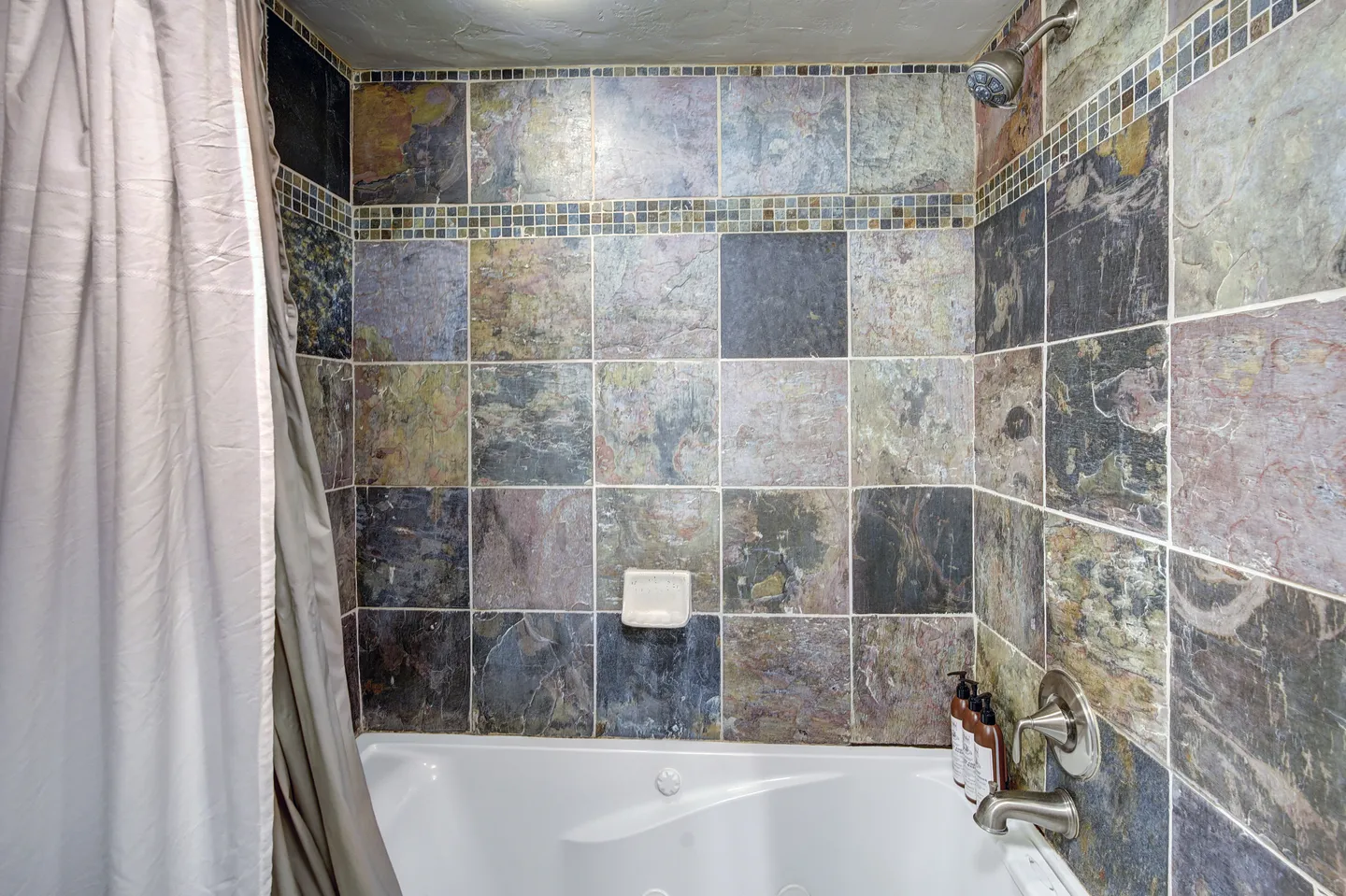 Bathroom shower and tub combination featuring rustic, multicolor stone-look tile with decorative mosaic borders and chrome fixtures.