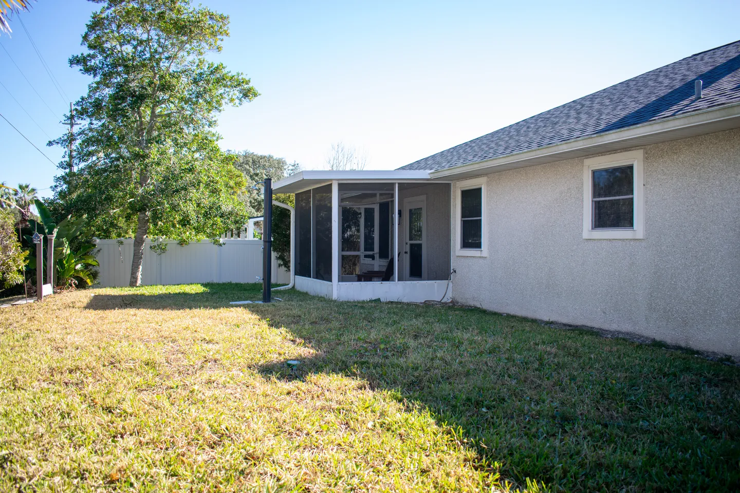 Rear exterior of a single-story house with a screened porch, grass yard, and white privacy fence under a clear blue sky.