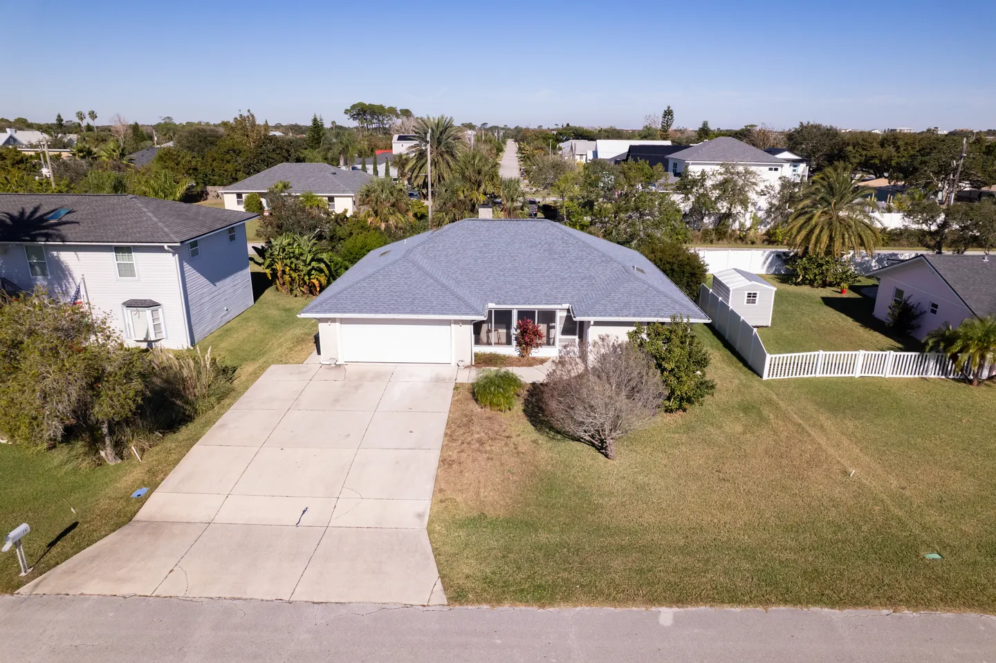 Aerial view of a single-story house with a grey roof, concrete driveway, and front lawn in a neighborhood.
