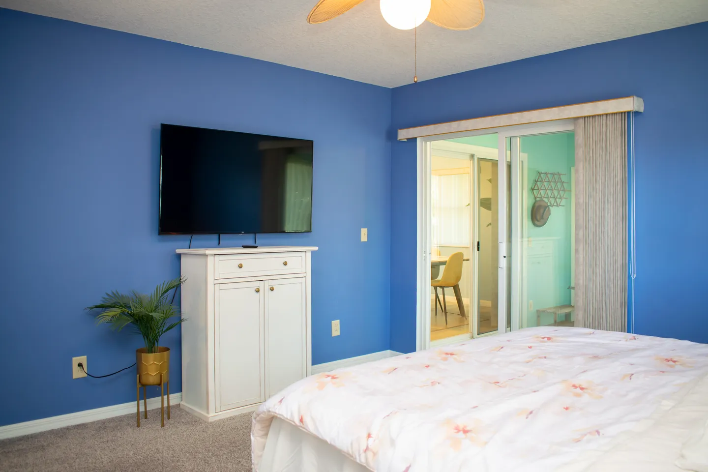 Bedroom with blue walls, carpeted floor, mounted TV, white cabinet, bed, and sliding glass doors.