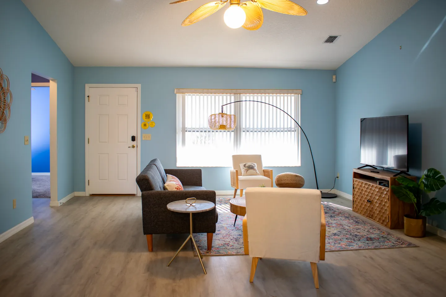 Living room with light blue walls, a dark grey sofa, light-colored armchairs, a television on a wooden console, and a window with vertical blinds.