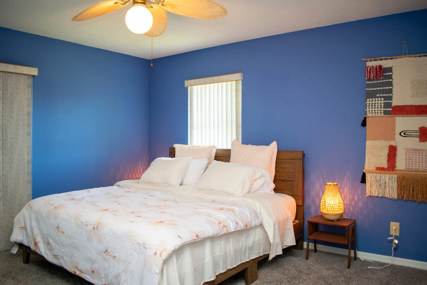 Bedroom with bold blue walls, a wooden bed with light-colored bedding, a nightstand with an illuminated lantern, and a woven tapestry.