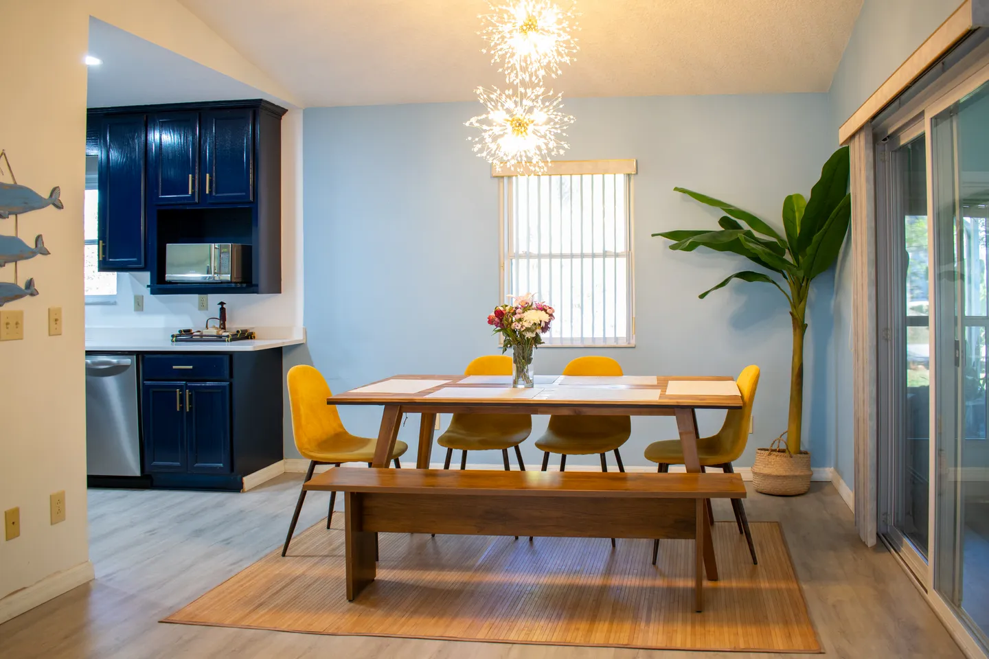 Dining area with a wooden table, bench, yellow chairs, and modern pendant lights adjacent to a kitchen with dark blue cabinets.