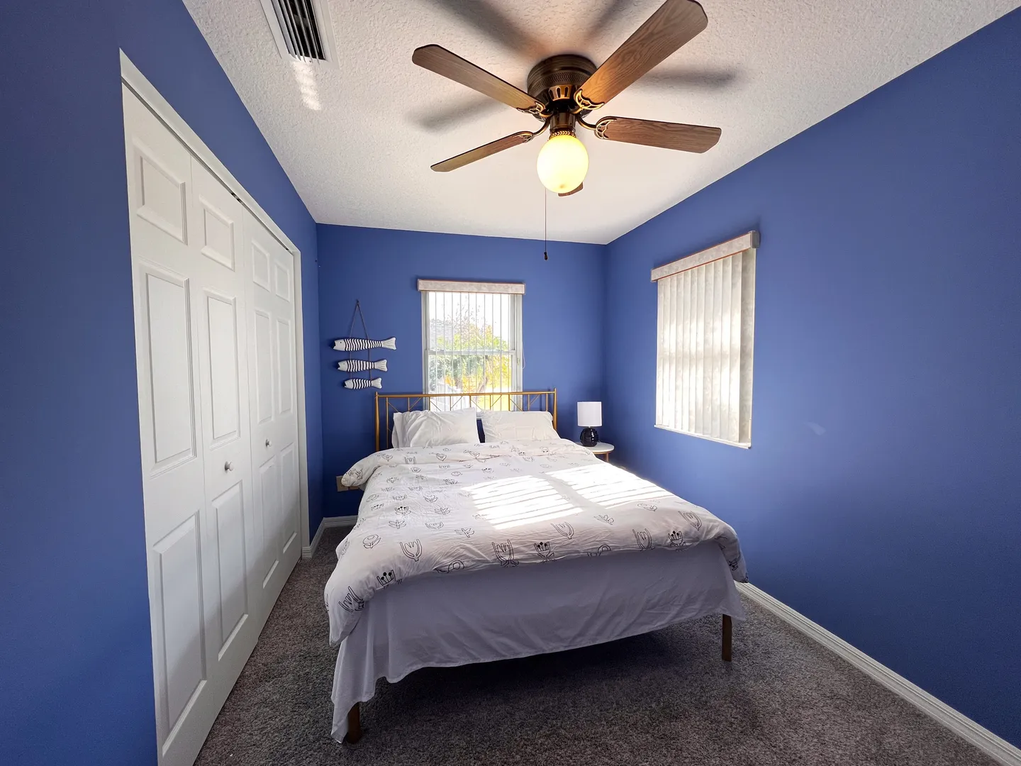 Bedroom with blue walls, carpeted floor, ceiling fan, bed with white bedding, two windows, and white bifold closet doors.