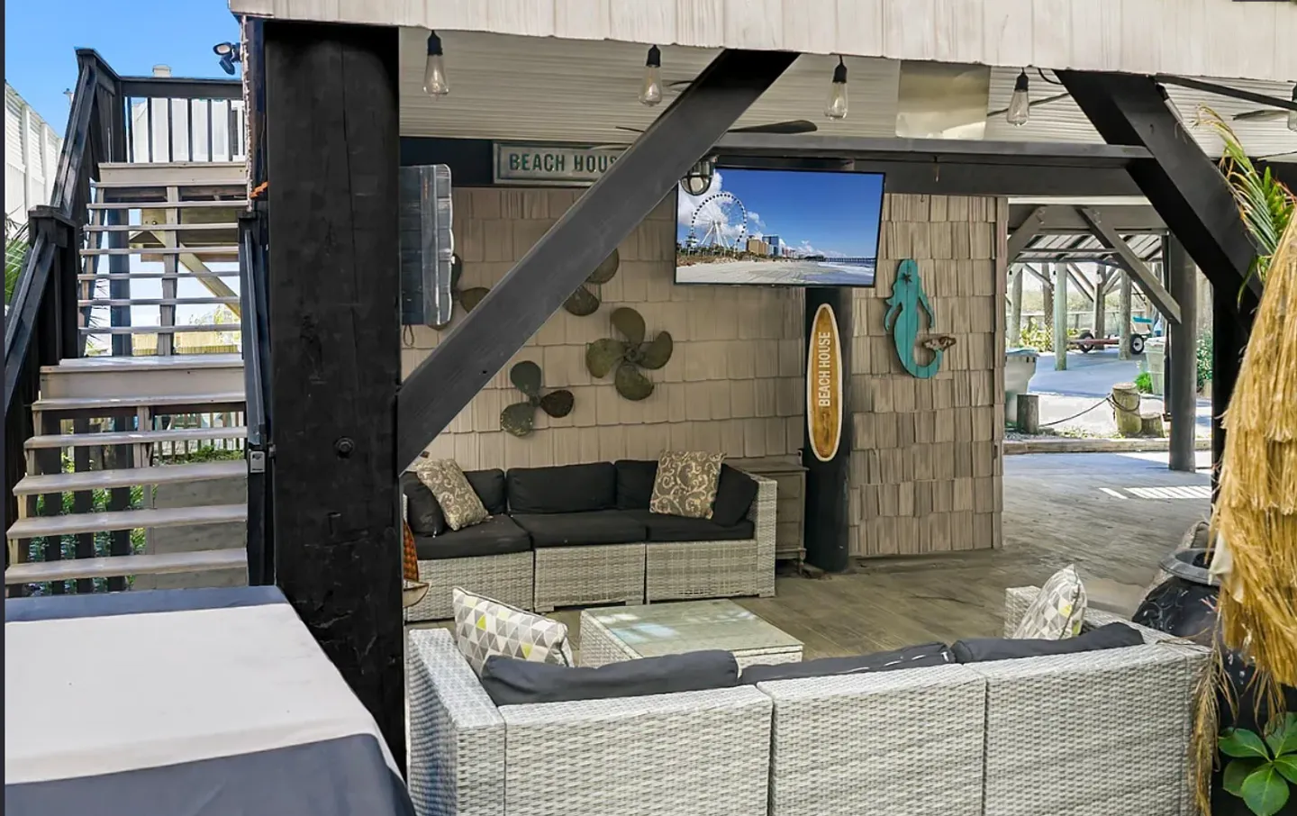 Covered outdoor patio beneath a stilt home featuring wicker sectional sofas, a coffee table, and a wall-mounted TV.
