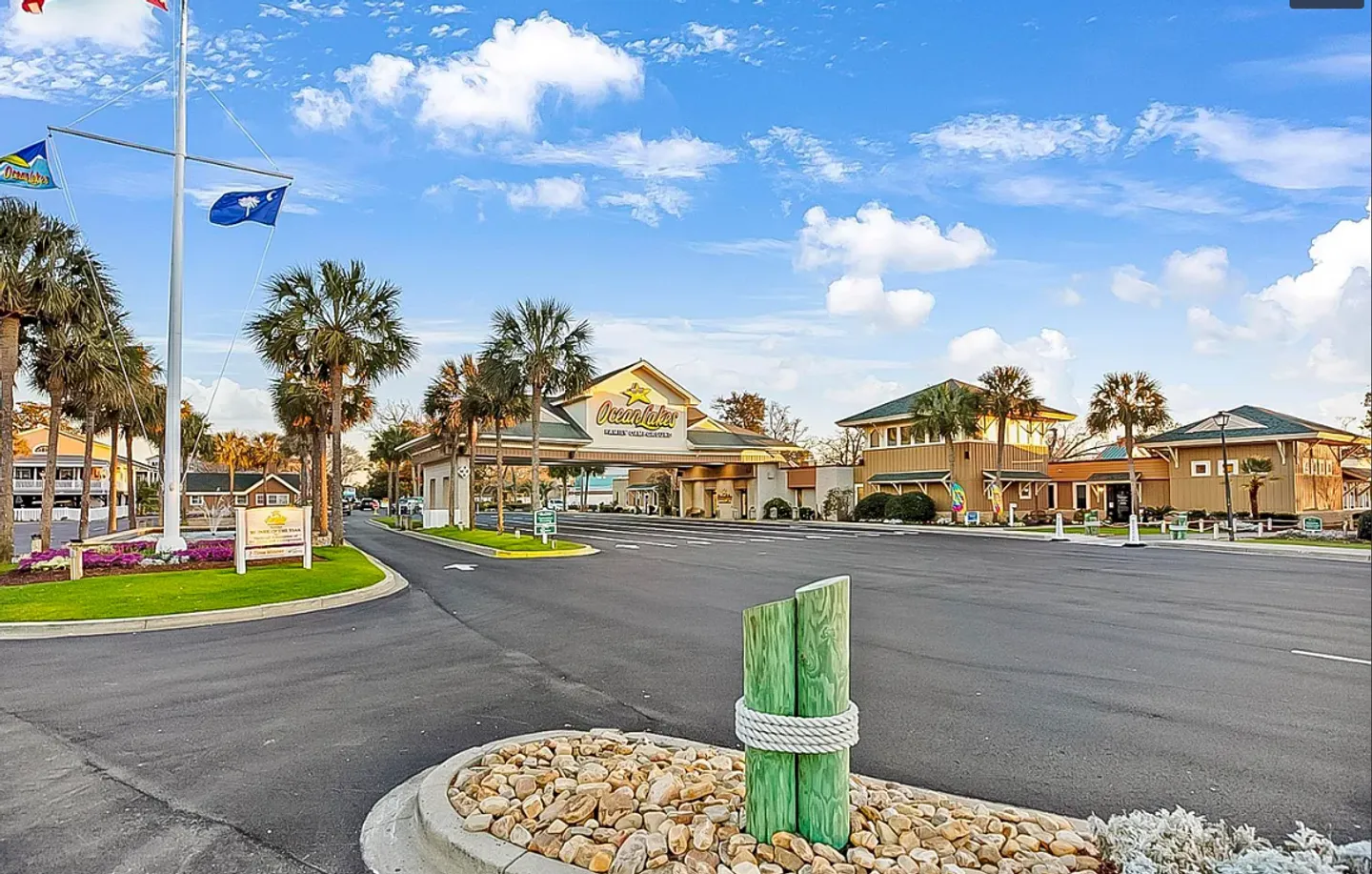 Paved entrance drive and gatehouse complex for Ocean Lakes Family Campground lined with palm trees under a blue sky.