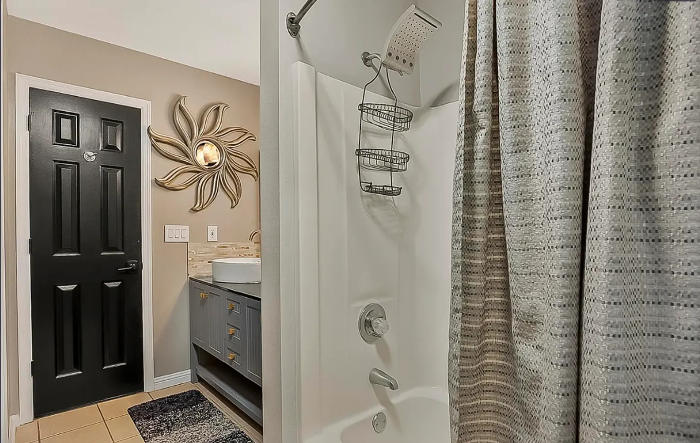 Bathroom showing a shower-tub combination in the foreground and a vanity with decorative mirror next to a black door in the background.