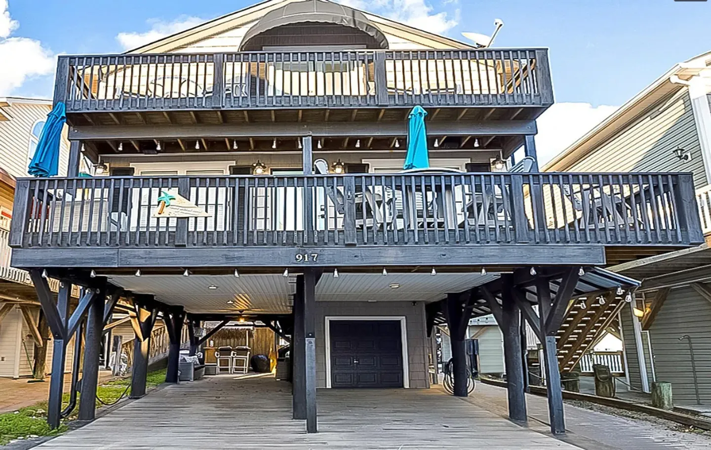 Exterior of a stilt house with multi-level dark wood decks, outdoor seating, and a ground-level covered carport.