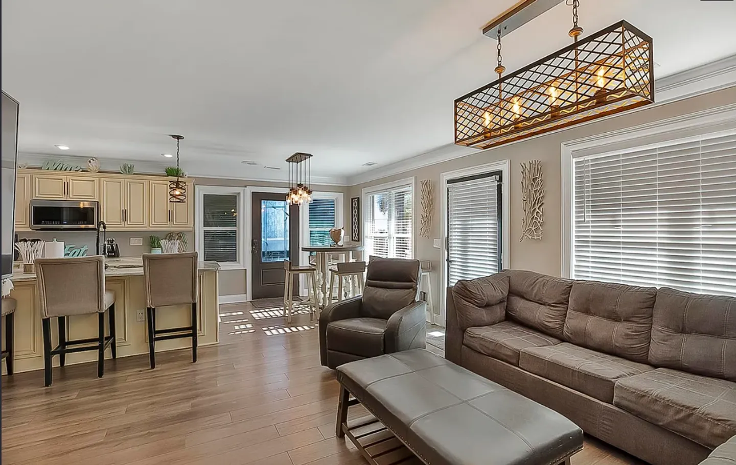 Open-concept living room and kitchen featuring a brown sofa, leather ottoman, kitchen island with bar stools, and wood-look flooring.
