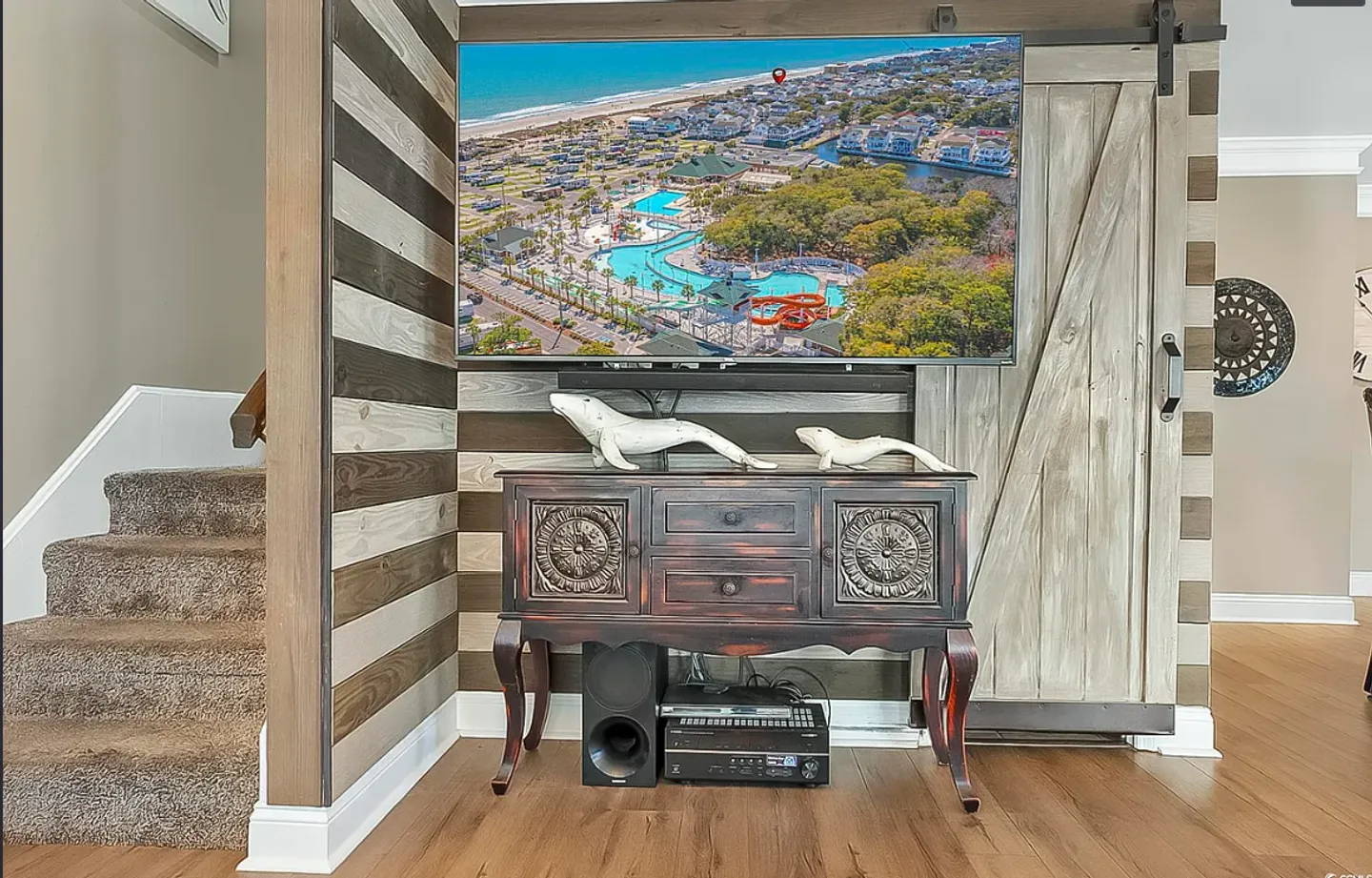 A wall-mounted TV displaying an aerial resort view above an ornate dark wood console table, set against a striped wood-paneled wall with a sliding barn door.