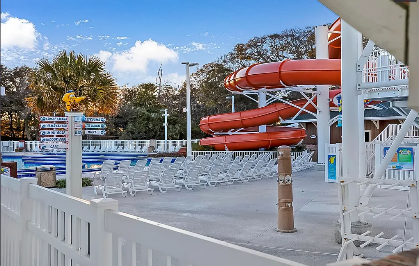 Outdoor resort pool area featuring a winding red waterslide, rows of white lounge chairs, directional signs, and palm trees under a blue sky.