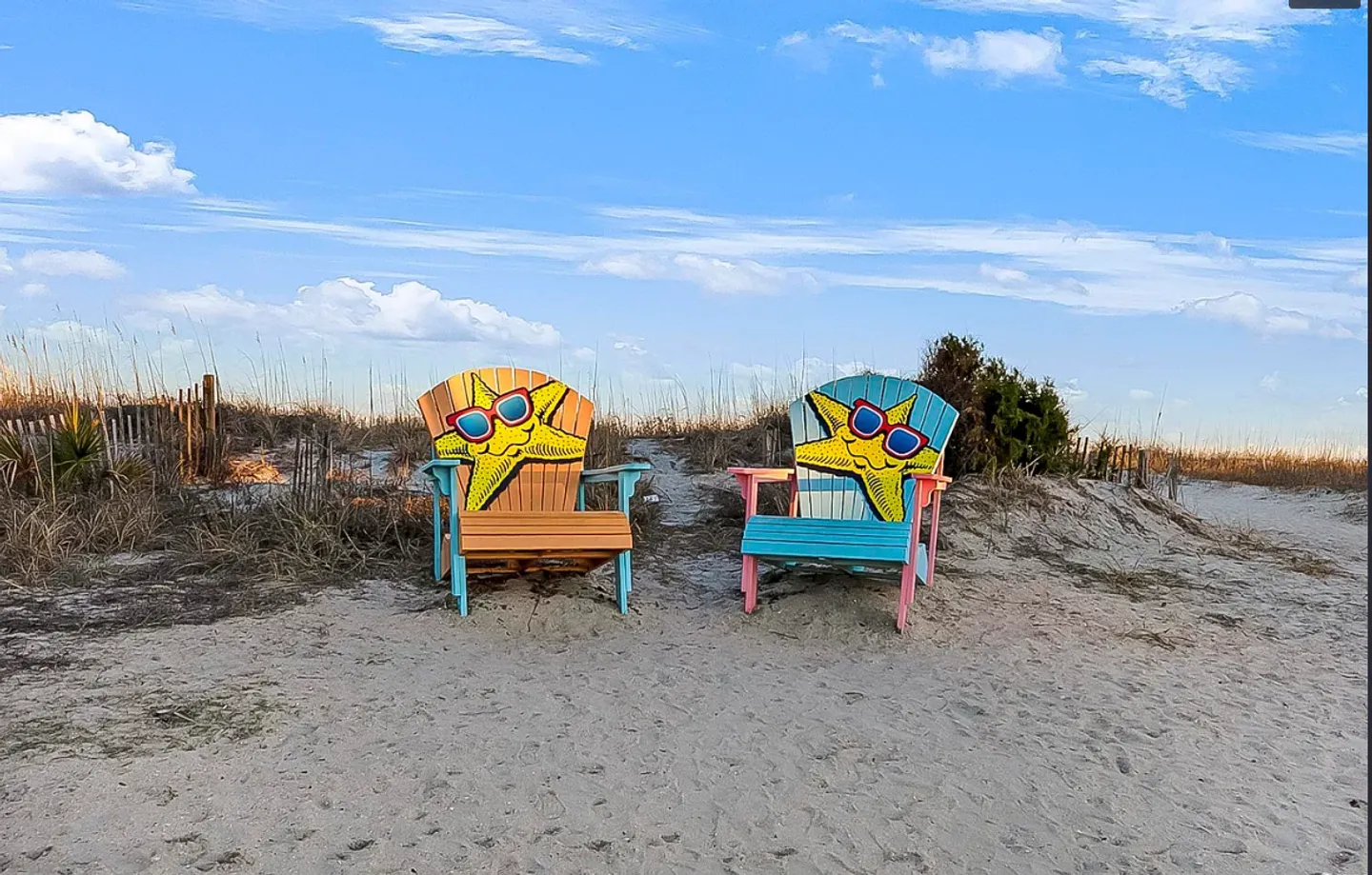 Two painted Adirondack chairs featuring cartoon starfish on the sandy beach path near sand dunes.