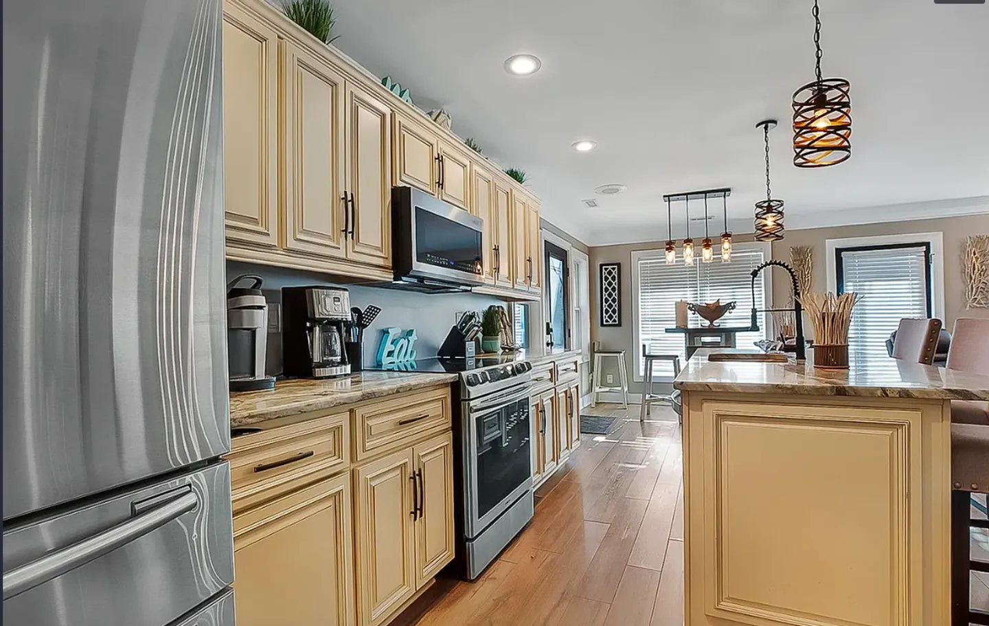 A kitchen with cream-colored cabinets, stainless steel appliances, a center island with pendant lighting, and wood-look flooring leading toward a dining area.