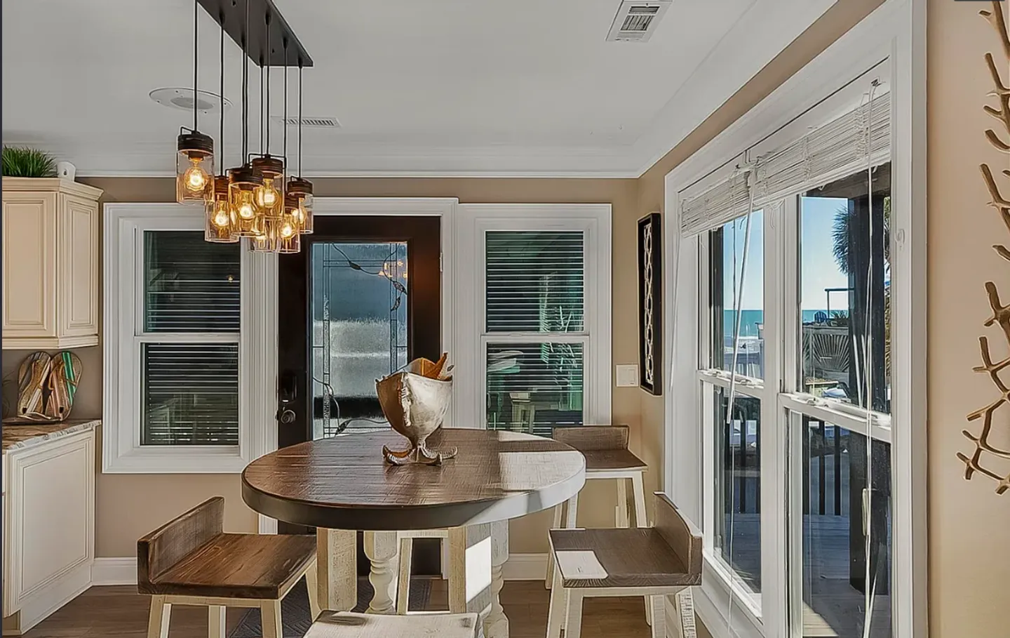 Dining area with a round wooden table, counter-height chairs, mason jar pendant lights, and windows overlooking the water.