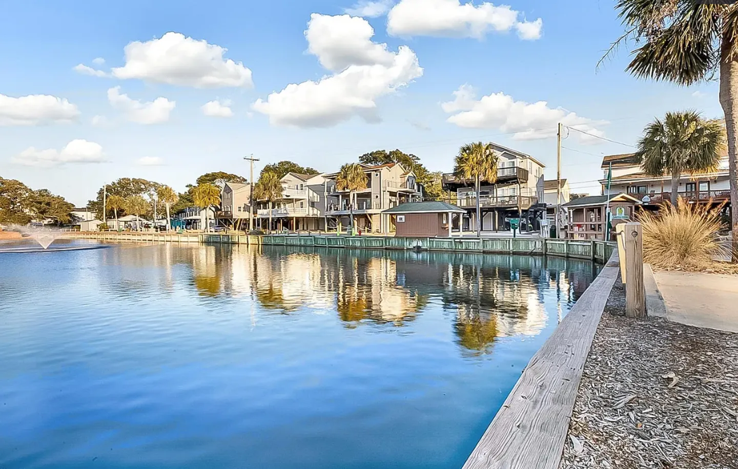 A wide view of a calm lagoon with a water fountain, bordered by a wooden edge, with multistory coastal homes and palm trees across the water under a blue sky.