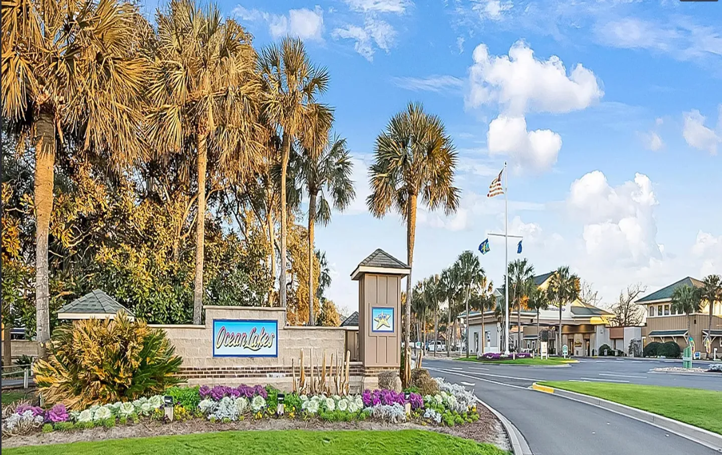 Entrance sign for Ocean Lakes with landscaping, palm trees, and an entry gate in the background under a blue sky.