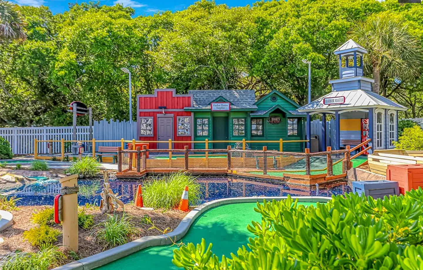 Outdoor miniature golf course featuring themed miniature buildings, a blue water hazard, and green turf surrounded by lush trees.