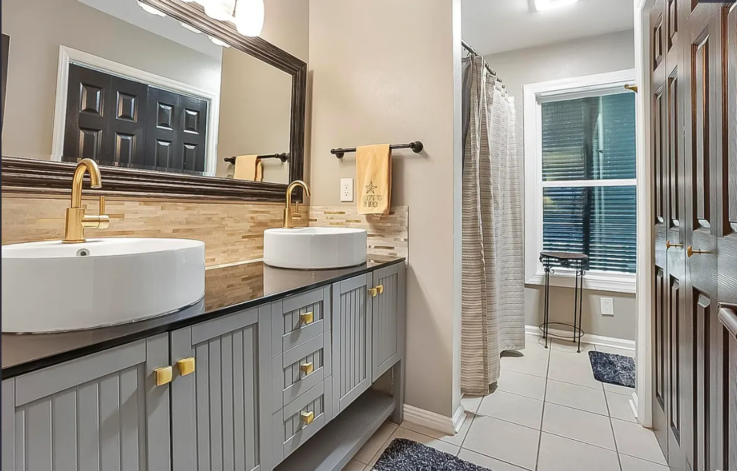 Bathroom with a double vanity featuring white vessel sinks, gold hardware, a large mirror, and tiled floors leading toward a shower curtain and window.
