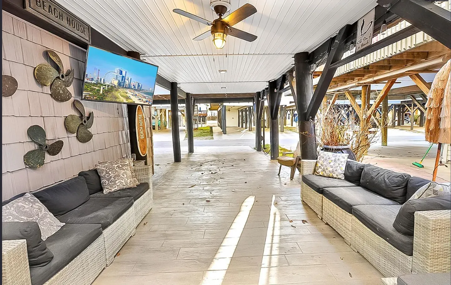 Covered ground-level patio beneath a stilt home, featuring outdoor sectional sofas, a mounted TV, and a ceiling fan.