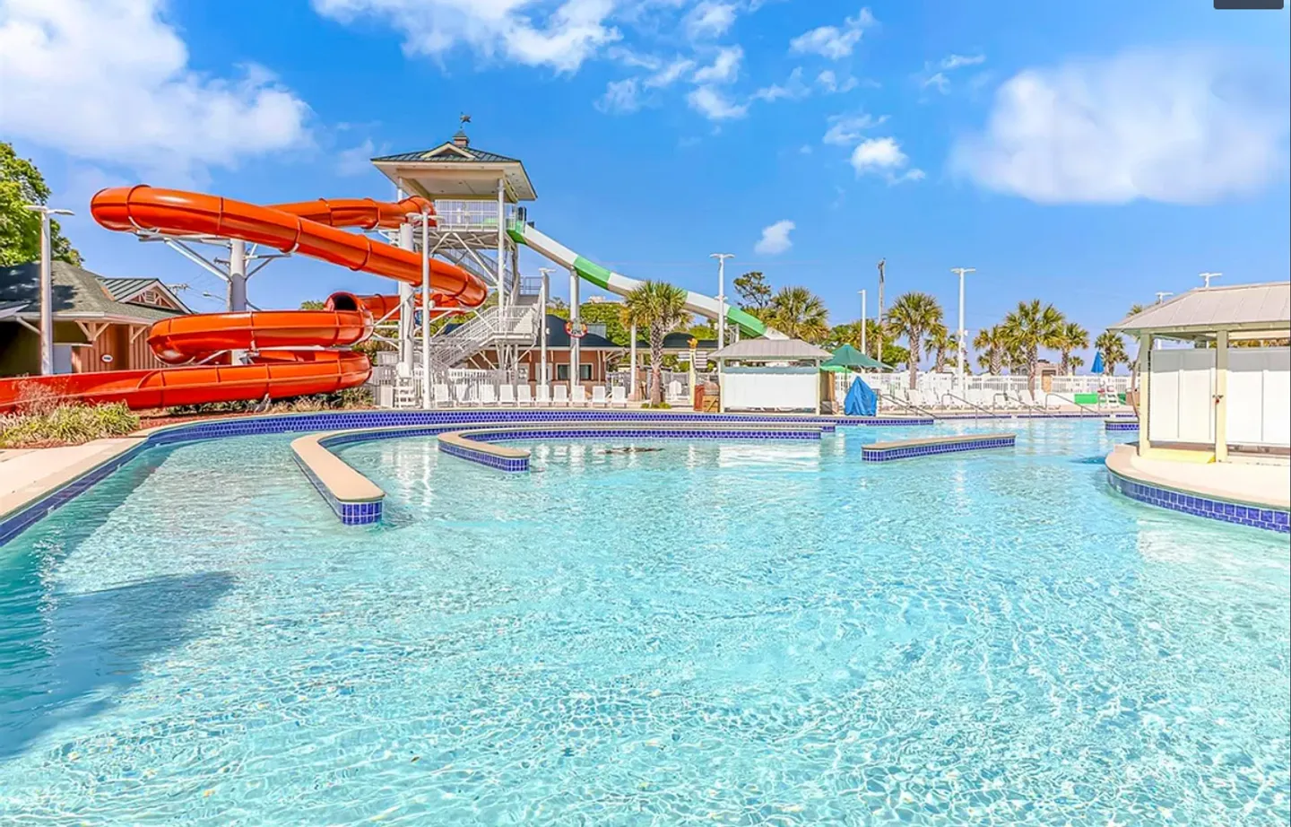 Outdoor resort pool complex with large water slides, poolside buildings, and palm trees under a bright blue sky.