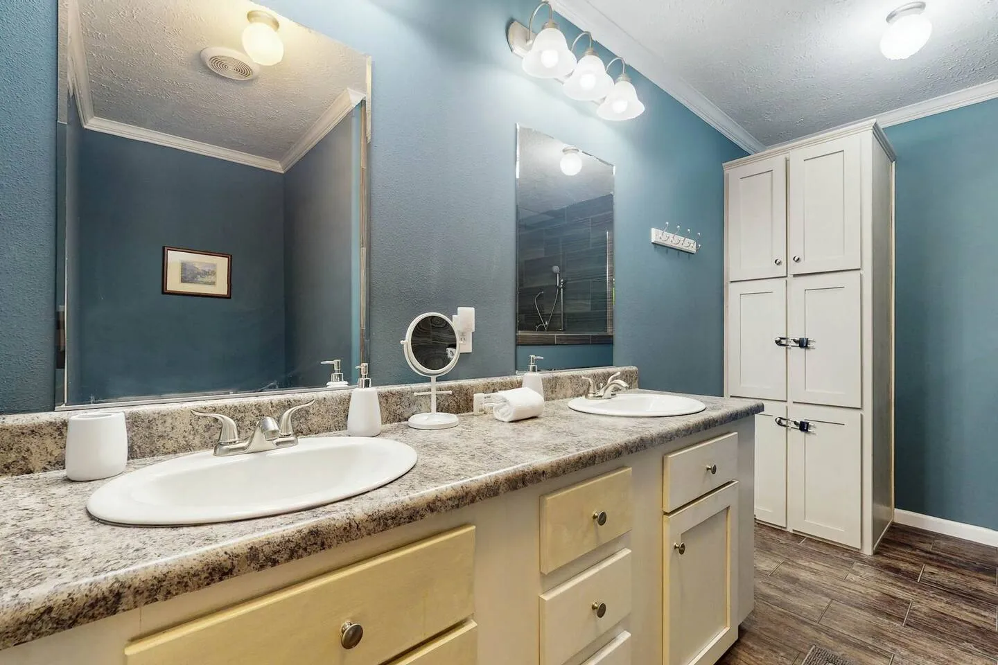Bathroom with a double sink vanity, two mirrors, blue walls, and a tall white cabinet.