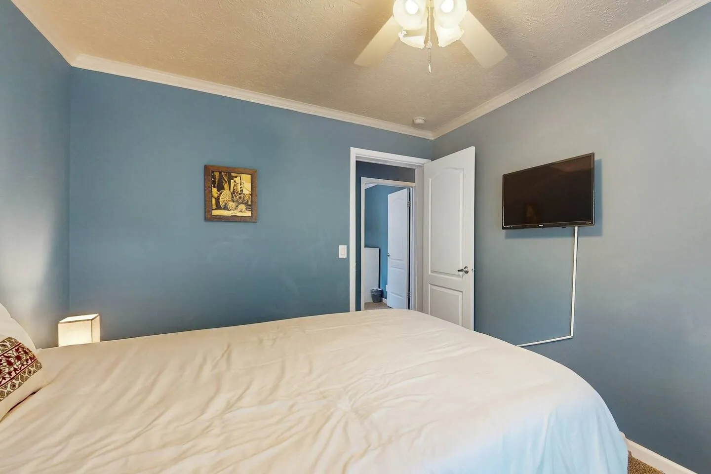 Bedroom with blue walls, a bed made with white linens, a ceiling fan, and a wall-mounted TV.