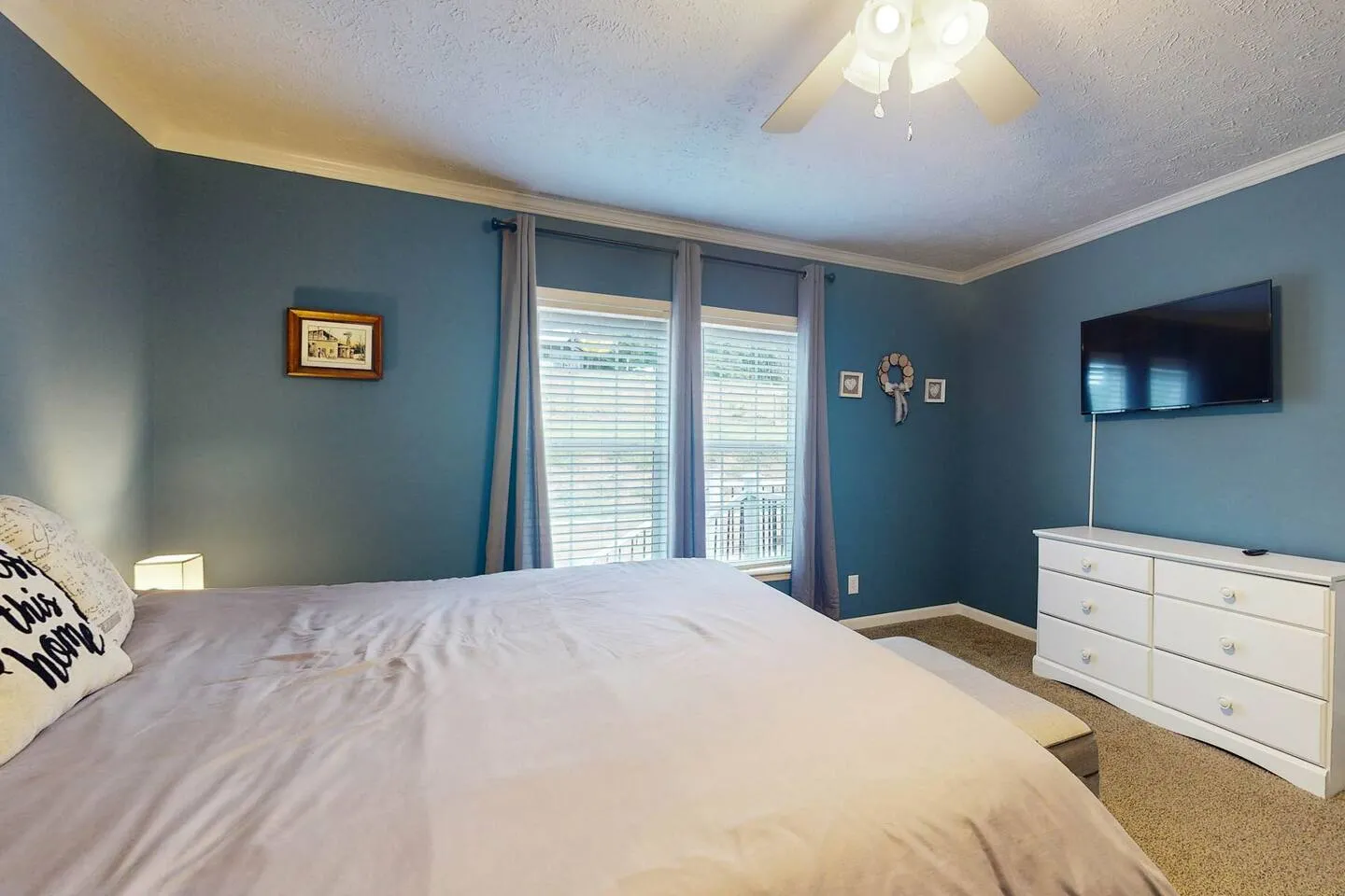 Bedroom with blue walls, carpet, a large bed with a light-colored duvet, two windows with blinds, a ceiling fan, a wall-mounted TV, and a white dresser.