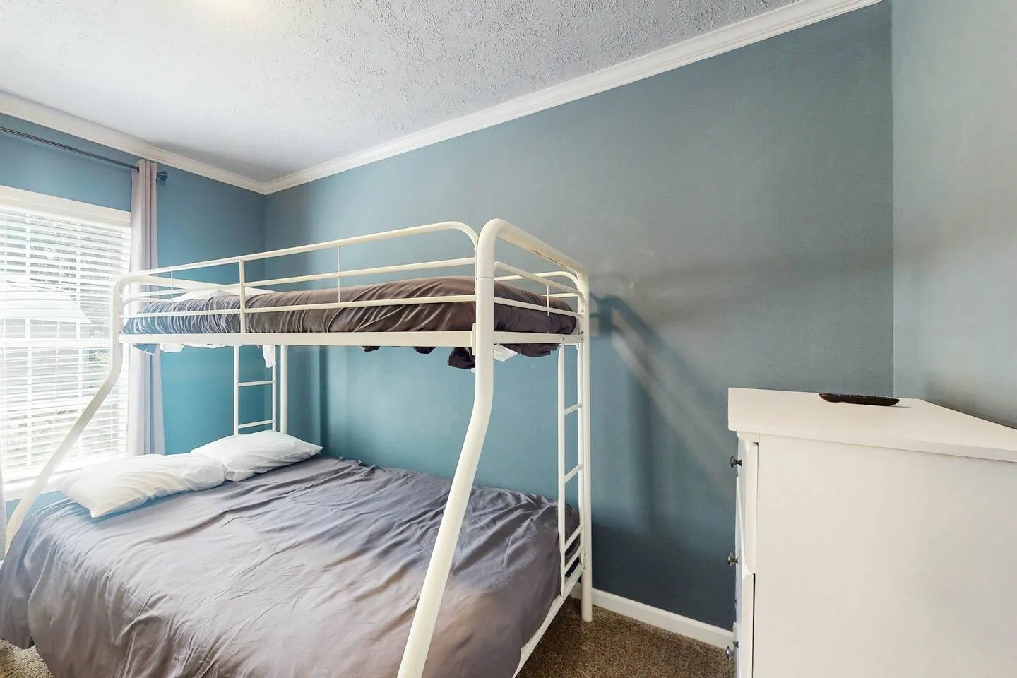 Bedroom with a white metal bunk bed against a light blue accent wall and a window with blinds.
