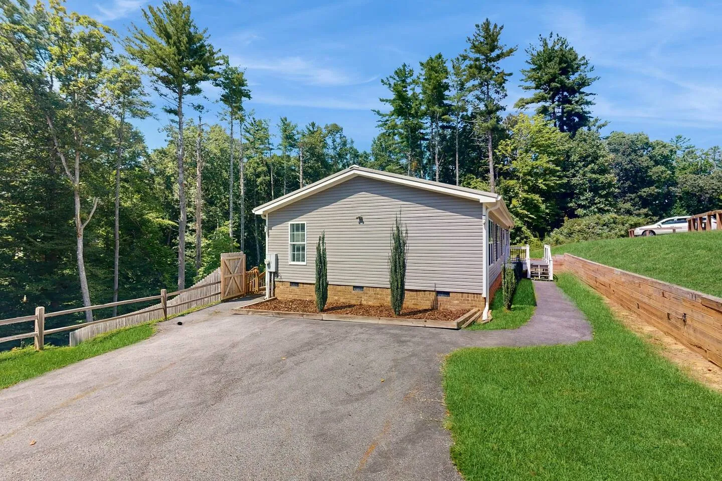 Exterior view of a single-story home with light siding, a paved driveway, and surrounding tall green trees.