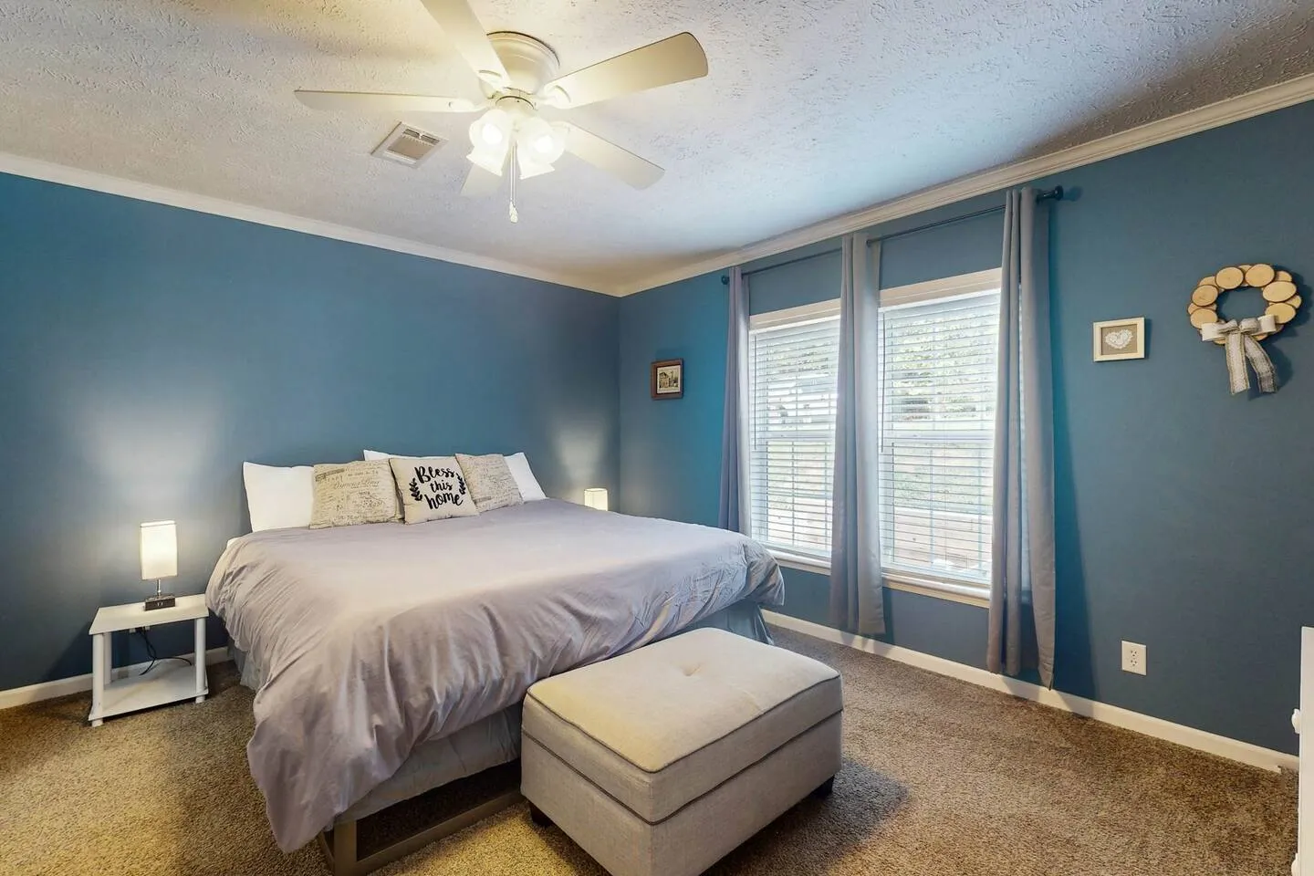 Bedroom with blue walls, carpeted floor, a bed made with gray bedding, an ottoman at the foot of the bed, and two windows with blinds and curtains.