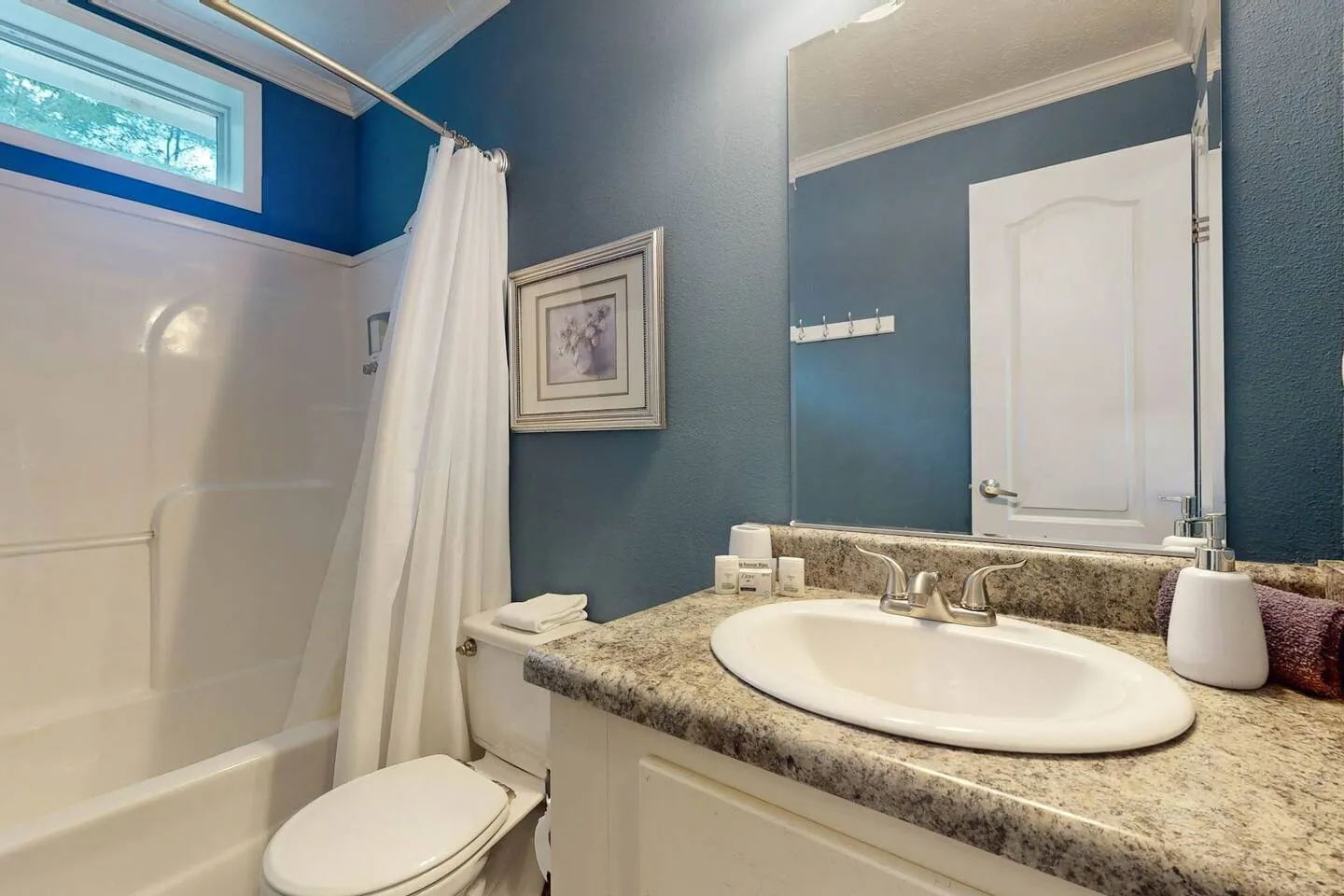 Bathroom with blue walls, a white vanity with granite-look counter, a mirror, toilet, and bathtub with a shower curtain.