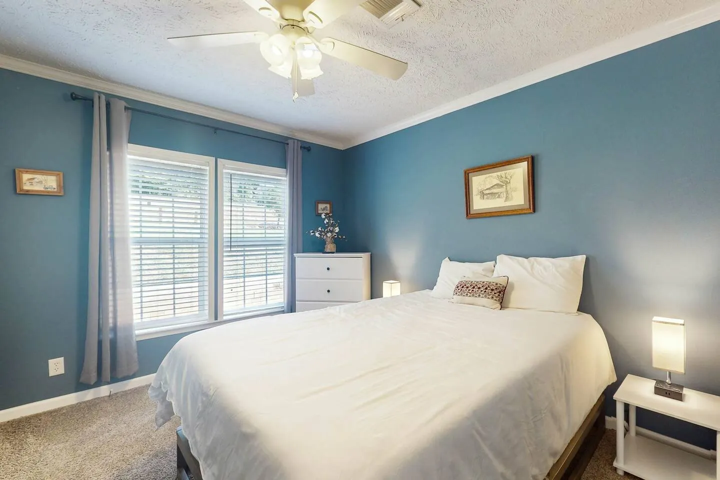 Bedroom with blue walls, carpeted flooring, a made bed with white linens, two windows with blinds, and a white ceiling fan.