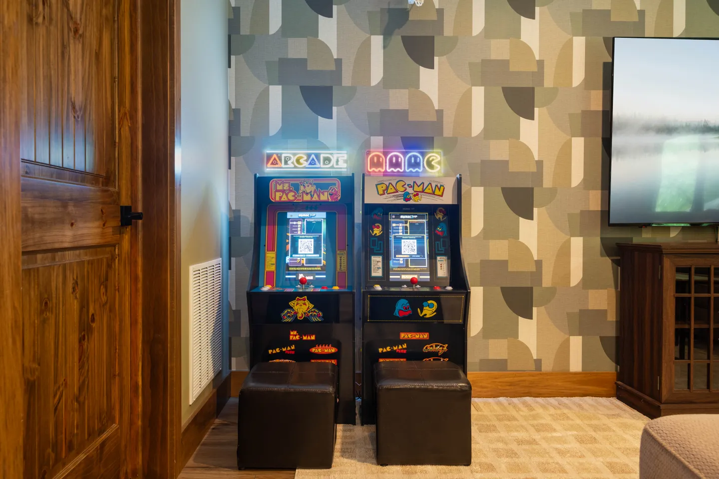 Two retro arcade cabinets with stools set against geometric wallpaper next to a television.