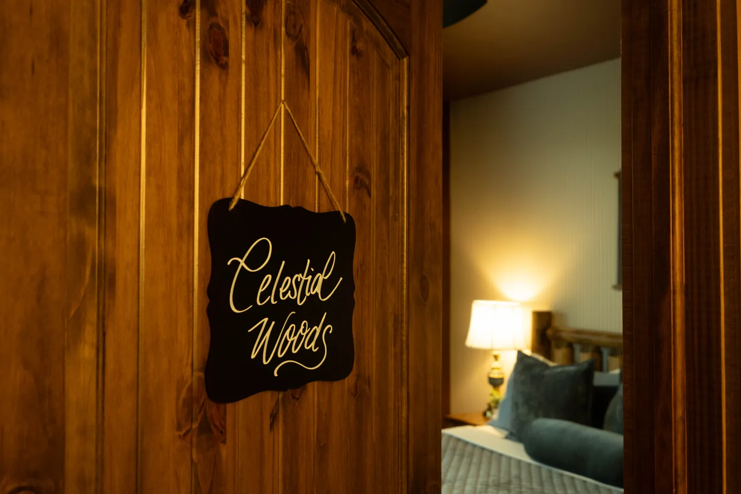 A wooden door with a hanging chalkboard sign reading 'Celestial Woods', opening into a warmly lit bedroom with a bed and lamp.