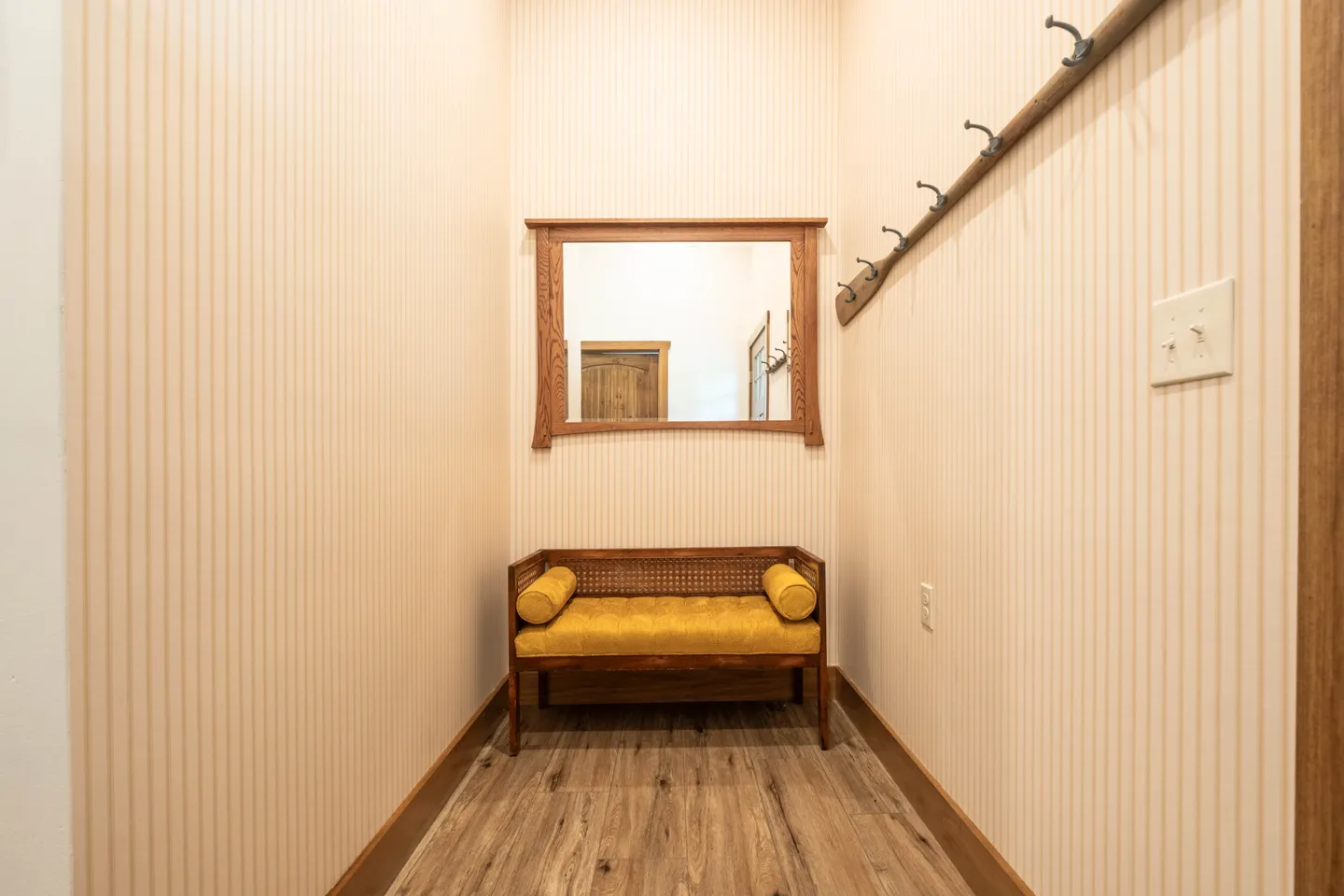 Narrow entryway hallway with striped wallpaper, wood-look flooring, a cushioned bench, a wood-framed mirror, and a wall-mounted coat rack.