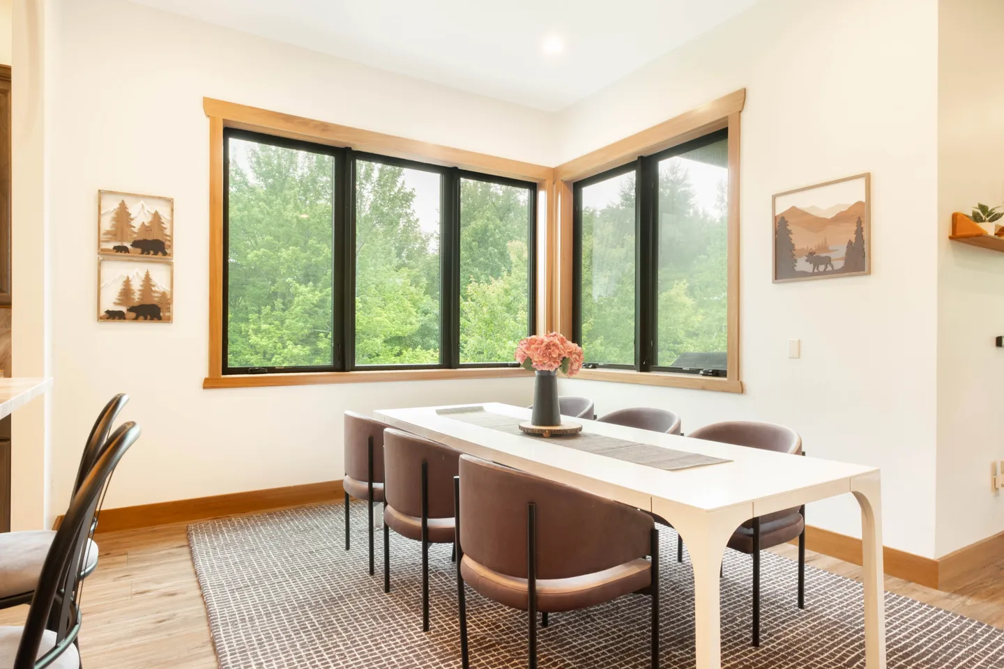 Dining area with a white rectangular table, modern rounded chairs, and corner windows looking out at green trees.