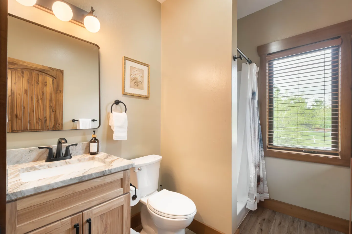 Bathroom with wooden vanity, mirror with vanity lighting, white toilet, and shower with curtain next to a window with blinds.