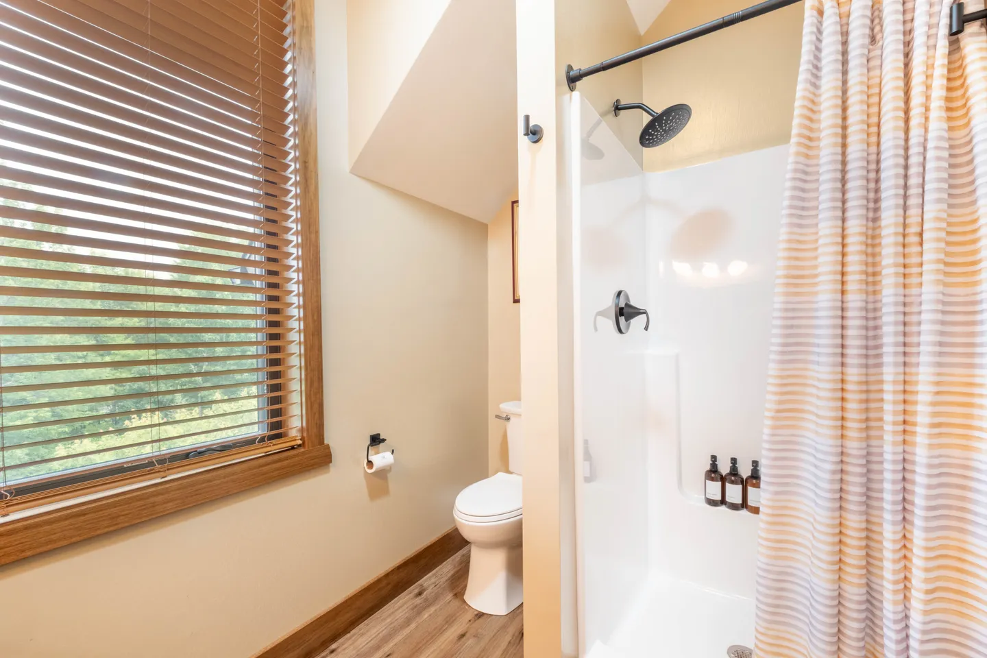 Bathroom showing a walk-in shower stall with a striped curtain, a white toilet under a sloped ceiling, and a window with blinds.