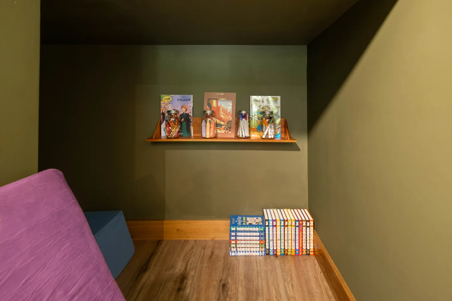A small alcove with olive green walls, wood-look flooring, a wall-mounted shelf with coloring supplies, and stacks of books on the floor.