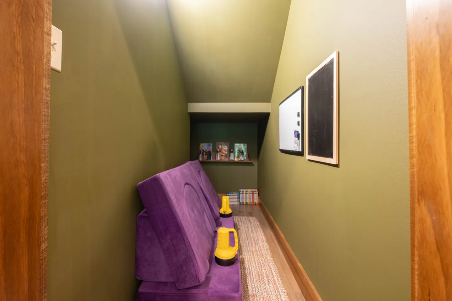 Cozy reading nook tucked under a staircase with olive green walls, purple floor cushions, yellow lanterns, and a dry erase board.