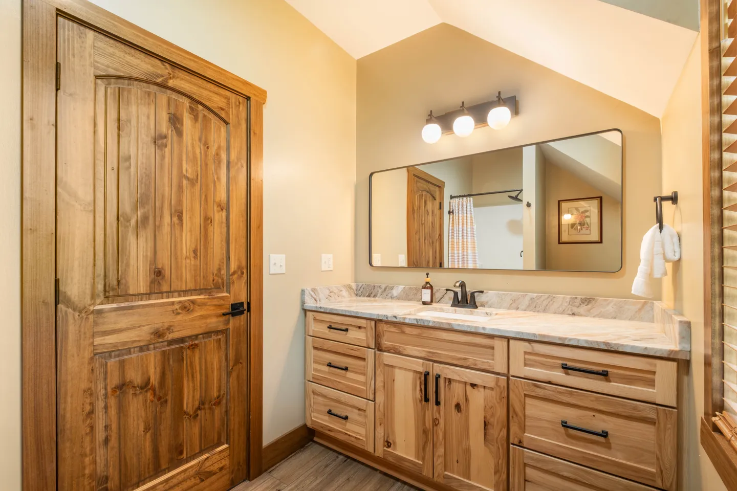 Bathroom with a natural wood vanity, marble-patterned countertop, wide rounded mirror, and matching solid wood door.