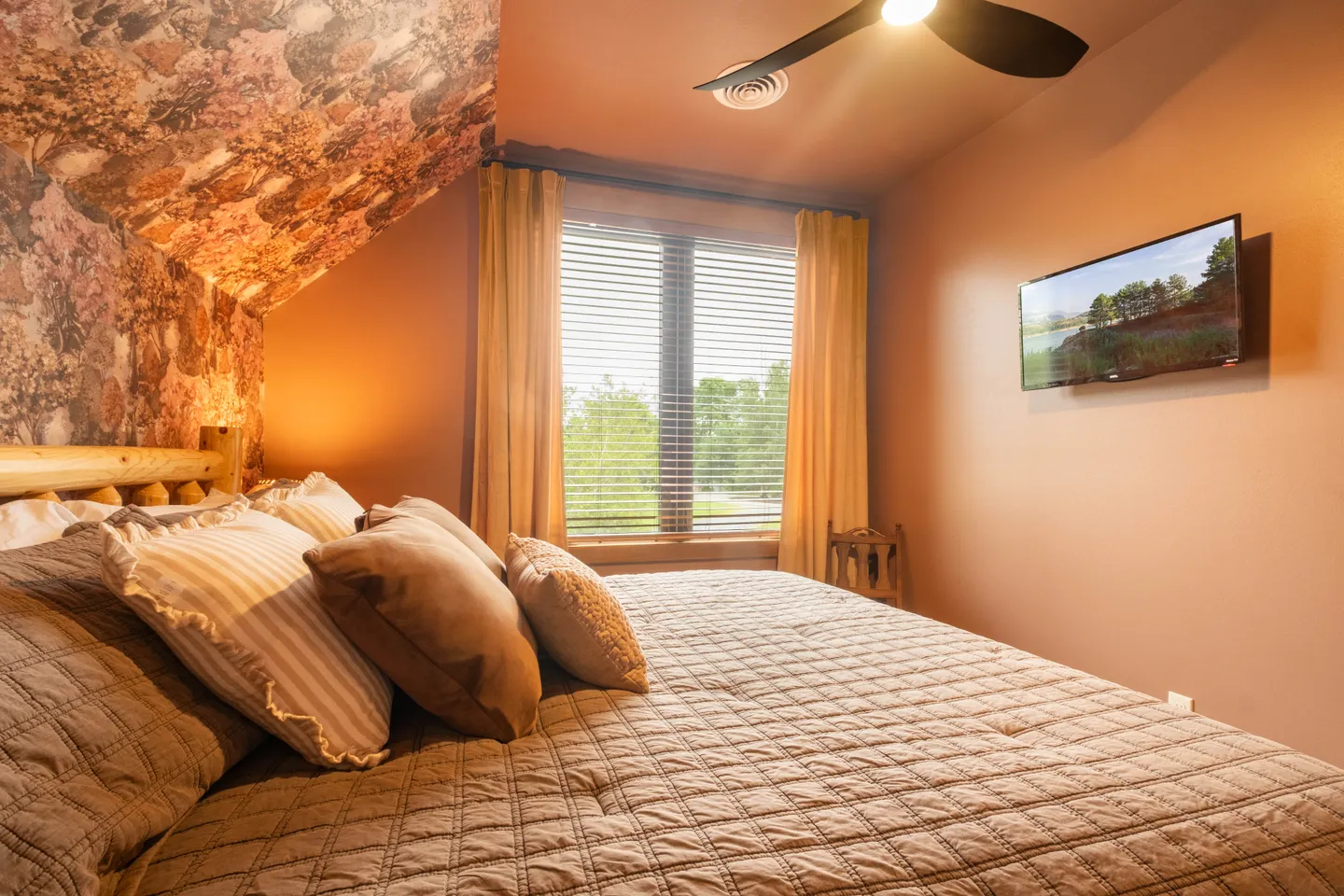 Bedroom with a bed, floral wallpaper on a sloped wall, a window with blinds and yellow curtains, and a wall-mounted TV.