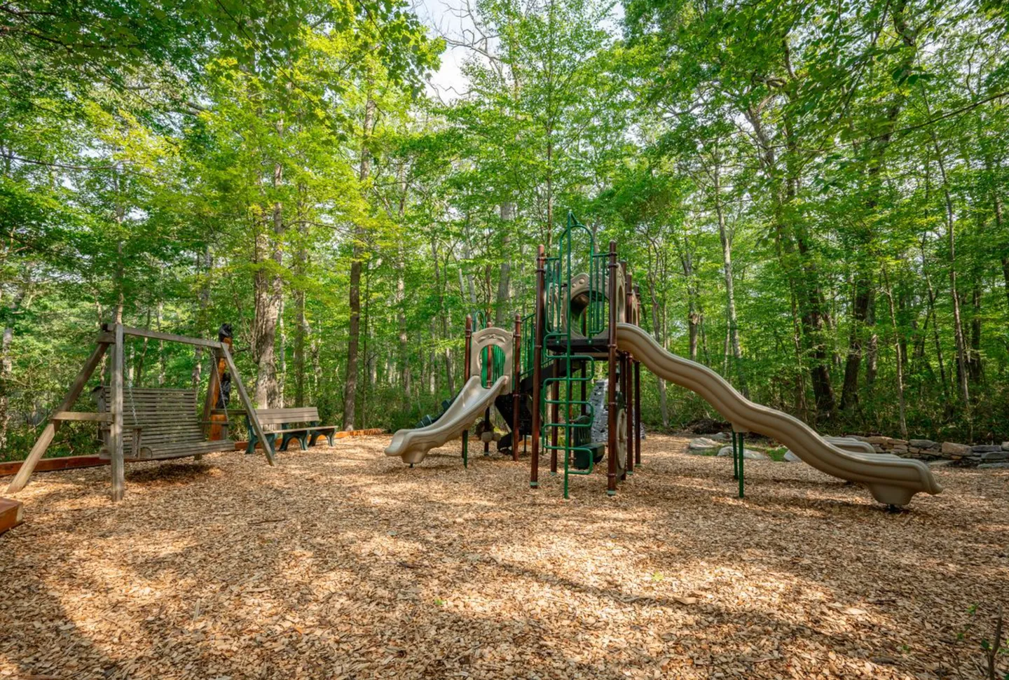 Outdoor playground playset with slides, a wooden swing, and a bench set over wood chips in a wooded area.