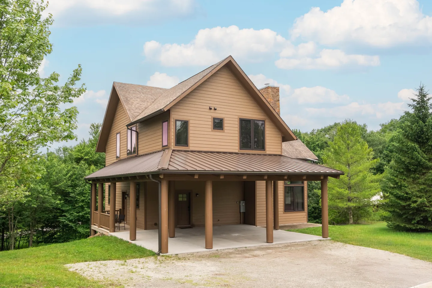 Exterior of a two-story brown house with a covered carport supported by round posts, surrounded by green lawn and trees.
