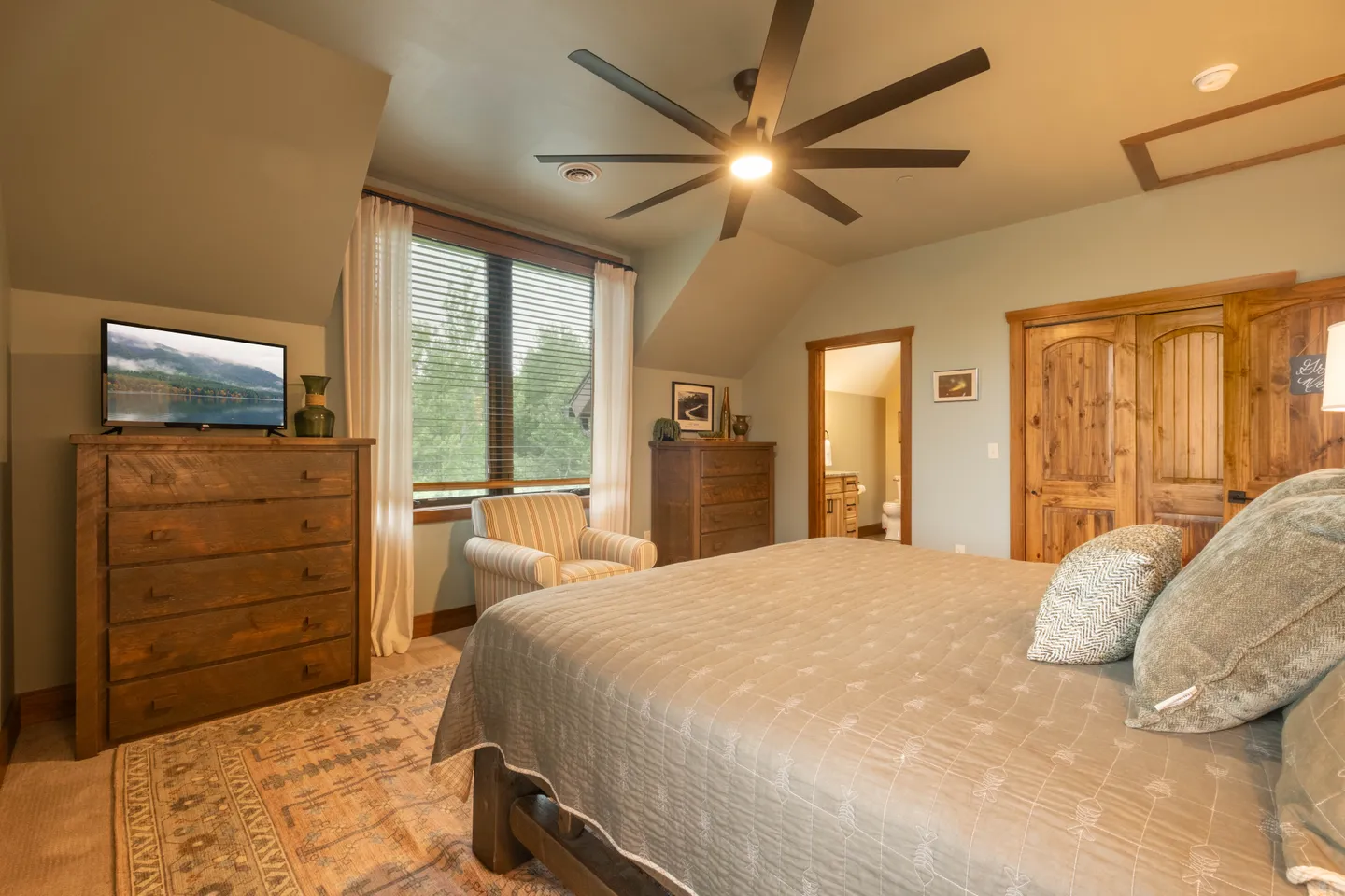 Bedroom featuring a made bed, patterned rug, wooden dressers, television, ceiling fan, and window looking out to trees.