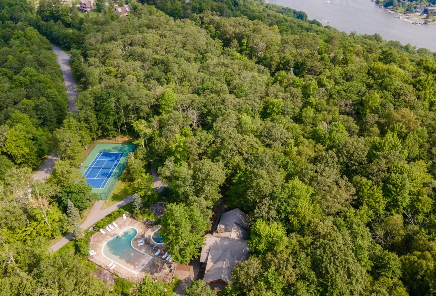 Aerial view of a wooded property showing a tennis court, swimming pool, and clubhouse nestled among dense green trees with a body of water in the background.