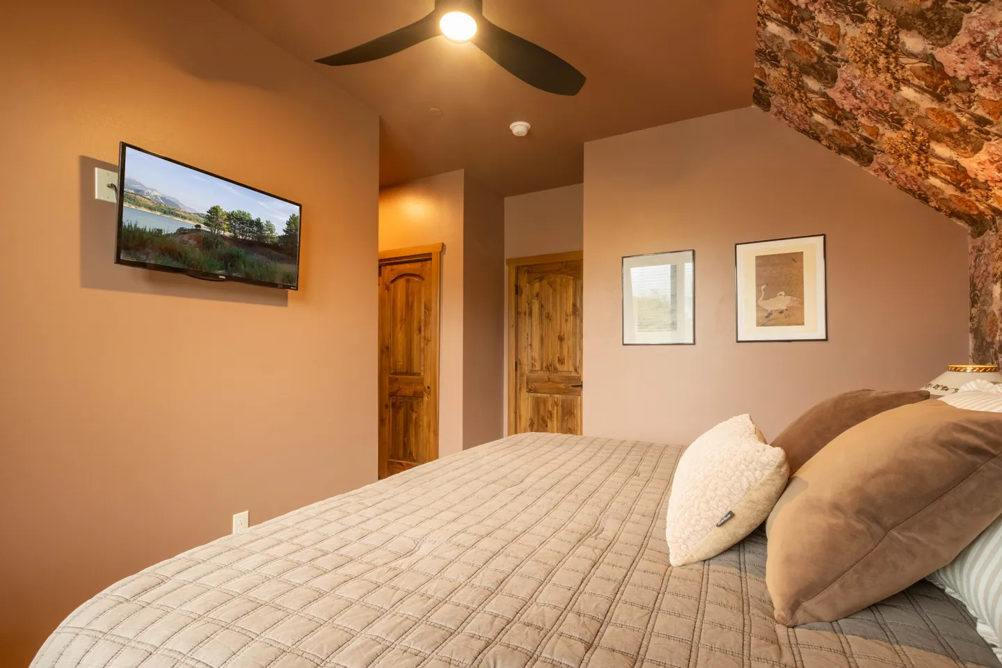 Bedroom with a quilted bed, wall-mounted television, ceiling fan with light, and rustic wooden doors.