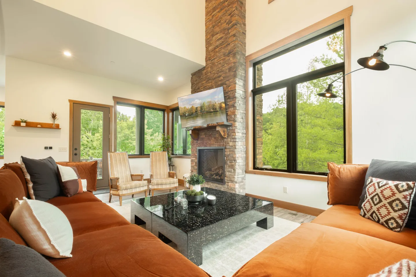 Bright living room with vaulted ceilings, large windows, a tall stone fireplace with a mounted TV, and rust-orange sectional seating.